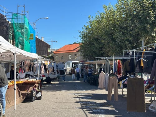 Escena de mercado al aire libre en Baiona con puestos de ropa, camiones de vendedores y edificios históricos bajo cielo azul despejado