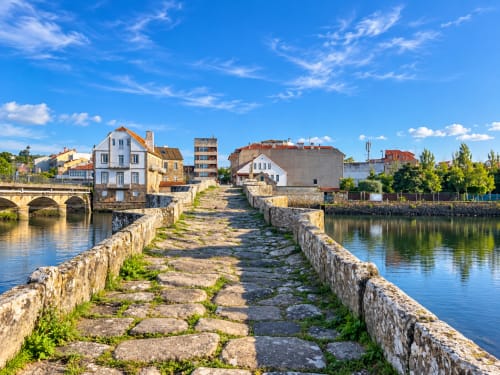 Antiguo muelle de piedra con vistas al frente marítimo de Baiona, edificios coloridos y cielo azul