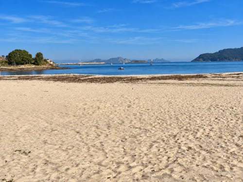 Playa arenosa del hotel Holibai en Baiona con mar azul cristalino e islas lejanas bajo cielos soleados