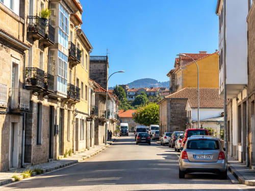 Calle histórica de Baiona con edificios de piedra, coches aparcados y vistas a las montañas de fondo