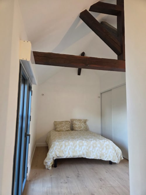 Dormitorio luminoso con vigas de madera oscura expuestas, cama con ropa de cama crema estampada, puerta azul y pisos de madera en La Perle du