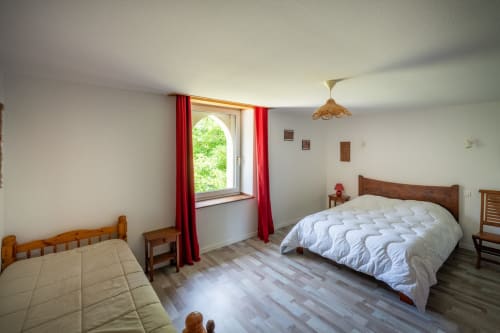 Chambre lumineuse au Domaine des Escouanes avec deux lits à cadre bois, fenêtre en arche aux rideaux rouges, et vue sur le jardin