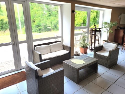 Lumineux salon-véranda du Domaine Des Escouanes avec sièges en rotin, vue sur le jardin par grandes fenêtres et plantes en pots.