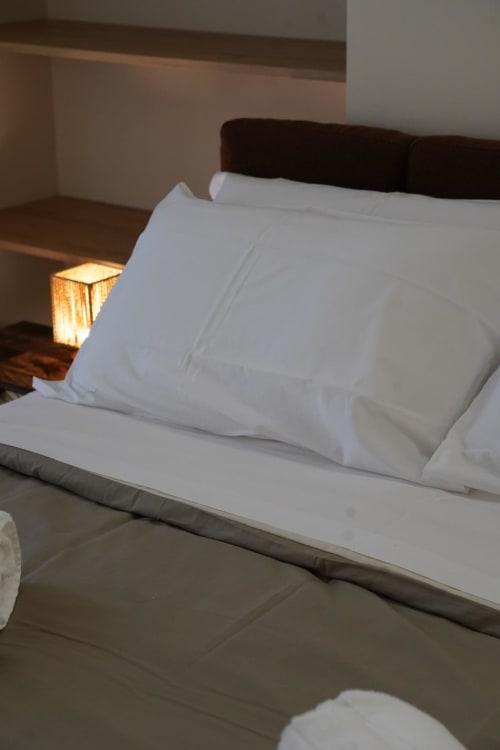 Camera da letto dell'hotel Domusè a Napoli con cuscini bianchi su testata scura e lampada da comodino calda