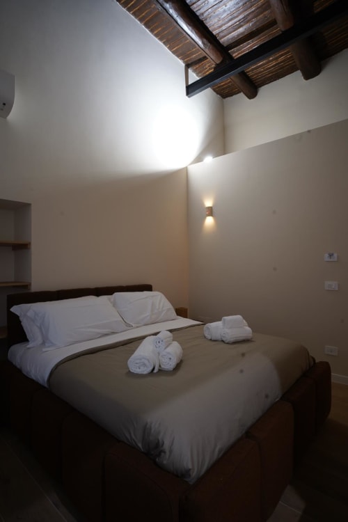 Camera moderna di Domusè Napoli con letto queen, biancheria bianca, asciugamani arrotolati, travi in legno, lucernario, stile minimalista