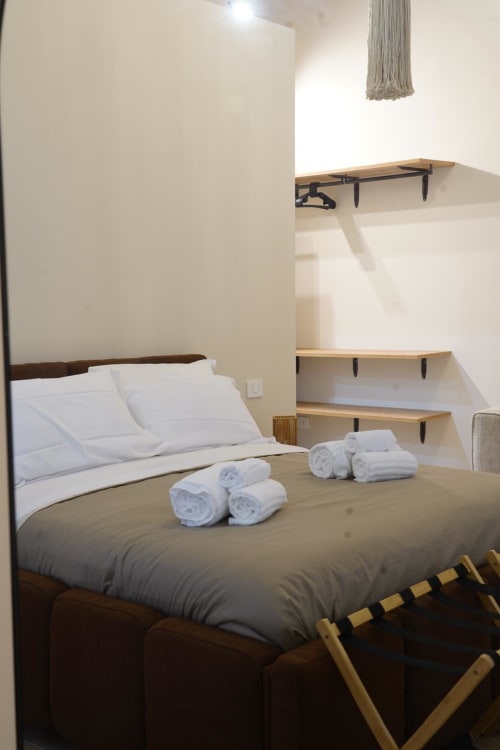 Camera San Gennaro a Domusè Napoli: letto con lenzuola bianche, asciugamani arrotolati, ripiani in legno e comfort moderni