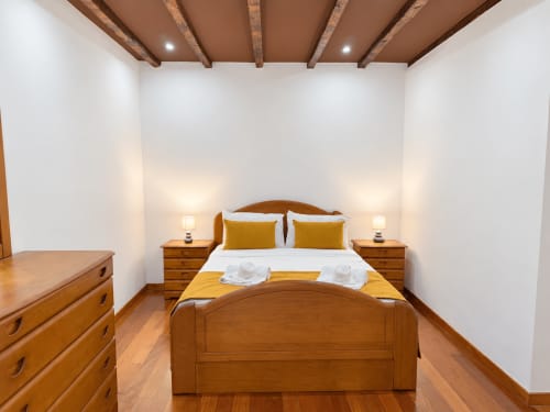 Wooden bedroom with exposed beams, queen bed with gold pillows, nightstands, and dresser at Check Madeira in Ribeira Brava