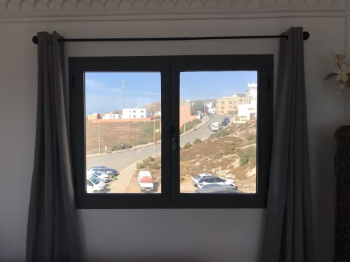 Black-framed window with black curtains overlooking Imsouane hillside with parked cars and colorful buildings