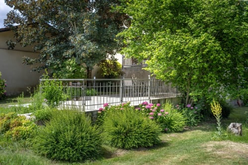 Jardin du Domaine Des Escouanes avec fleurs roses, arbustes verts et passerelle blanche en métal au-dessus de la terrasse