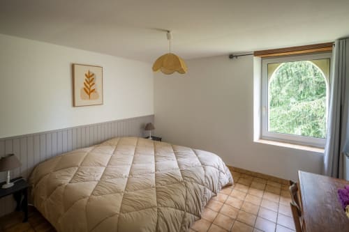 Burley cottage bedroom with garden view at Domaine Des Escouanes, featuring cream bedding, wooden pendant lamp, and