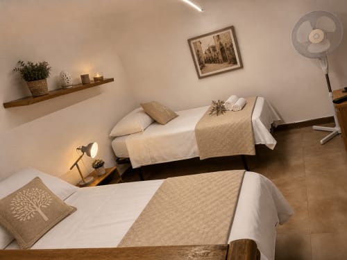 Letti gemelli con coperte beige nell'appartamento di Terlizzi. Mensola in legno, lampada da tavolo, paesaggio urbano incorniciato e ventilatore.