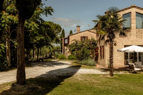 Hotel in laterizio toscano con viale alberato, giardini curati e ombrelloni bianchi a Buonconvento