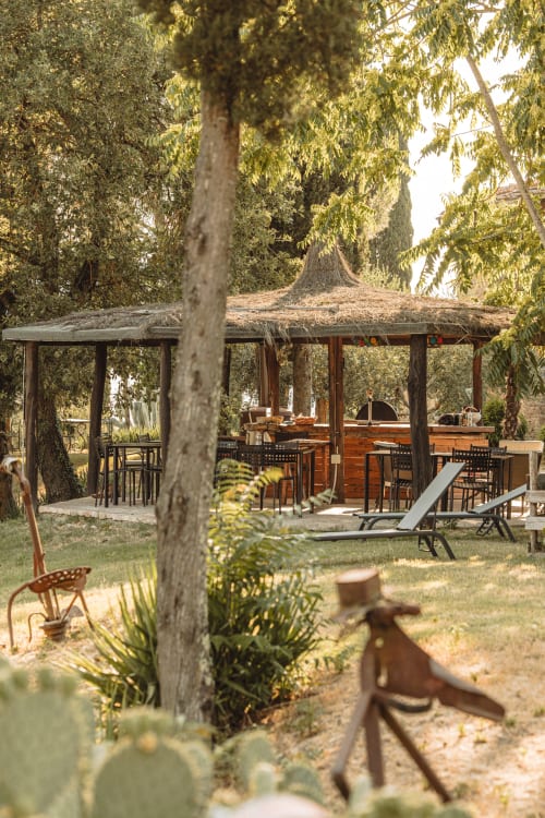 Giardino di hotel toscano con padiglione in paglia, bar in legno, zona ristorazione e lettini circondati da cipressi e ulivi