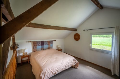 Chambre lumineuse sous les combles avec poutres apparentes, literie rose et vue sur le jardin du gîte Domaine Des Escouanes