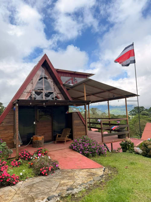 Chalets Vistas del Poas villa in Heredia with red-roofed A-frame structure, Costa Rican flag, mountain views, and flowering