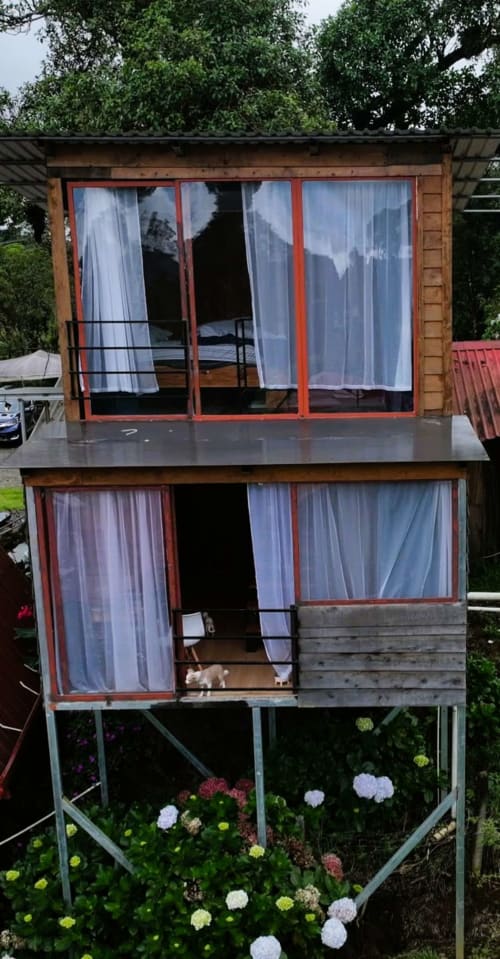 Cabaña de madera de dos pisos en Chalets Vistas del Poás, Heredia, con ventanas de marco rojo, cortinas blancas y flores de colores