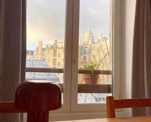 Vue depuis une fenêtre d'appartement parisien sur la basilique du Sacré-Cœur et les toits de Montmartre, avec rideaux rayés et plante en pot