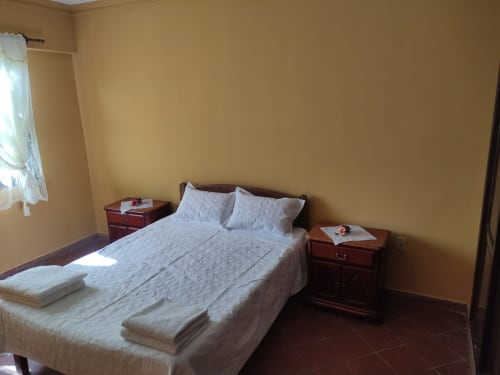Dormitorio limpio en apartamento de Tarija con cama queen, ropa de cama blanca, mesitas de madera y paredes amarillas