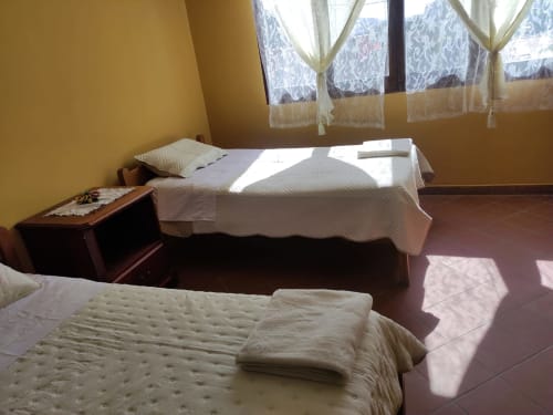 Dormitorio luminoso en Tarija con dos camas, paredes amarillas, cortinas de encaje, mesita de noche de madera y piso de baldosas