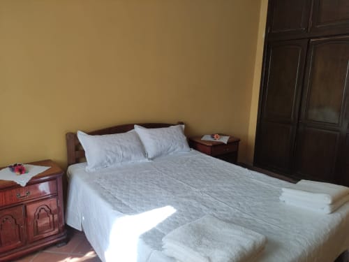 Dormitorio luminoso en apartamento de Tarija con ropa de cama blanca, edredón gris, mesitas de madera, armario oscuro y paredes amarillas