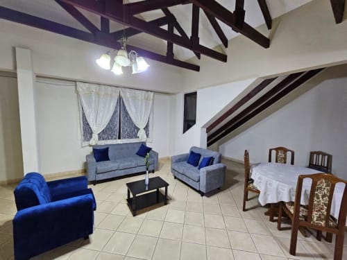 Amplia sala de estar en apartamento de Tarija con asientos azules, comedor, vigas expuestas y vistas a la ciudad desde las ventanas