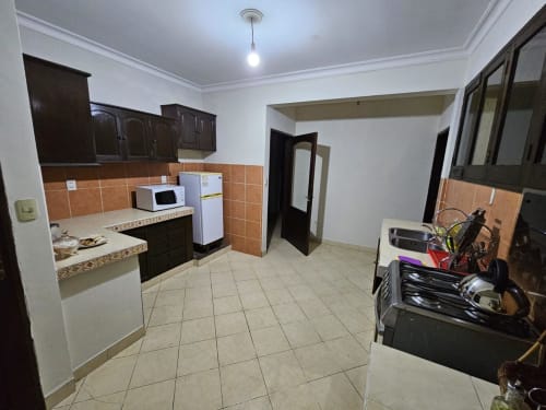 Amplia zona de cocina-sala en apartamento de Tarija con muebles de madera oscura, backsplash de azulejos naranjas, electrodomésticos blancos y pisos de azulejo