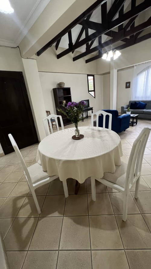 Comedor con mesa redonda blanca y sillas blancas en apartamento de Tarija, con vigas negras expuestas y flores púrpuras