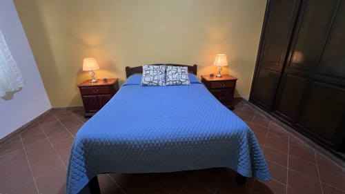 Dormitorio en apartamento de Tarija con cama acolchada azul, mesitas de noche en madera oscura, iluminación cálida y piso de baldosas de terracota