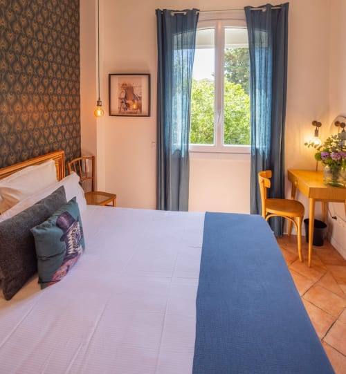 Chambre double lumineuse avec literie blanche, plaid bleu, mobilier en bois, papier peint à motifs et vue sur le jardin