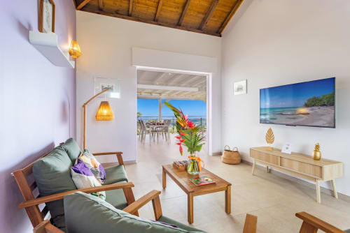 Salon de villa moderne avec vue sur mer, fleurs tropicales, mobilier en bois, téléviseur écran plat et terrasse vue océan