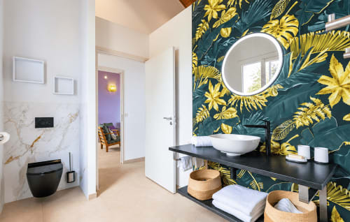 Salle de bain moderne avec plan de travail noir, vasque blanche ronde, miroir circulaire et papier peint bleu canard aux motifs botaniques jaunes