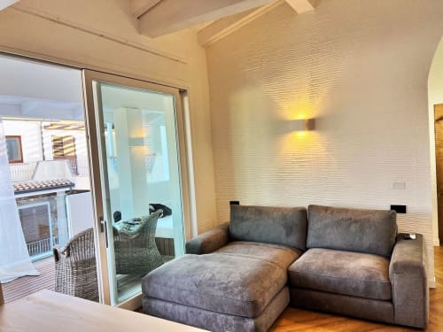 Salotto moderno con divano grigio componibile, pareti bianche strutturate e porte vetrate che si aprono sul balcone giardino di Palau