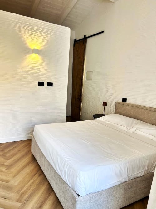 Camera moderna a La Sciumara Resort Palau con letto beige, pareti bianche texturizzate, porta a soffitta in legno e pavimento in legno duro