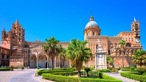 Cattedrale di Palermo con architettura in terracotta, cupola, palme e giardini curati sotto cielo azzurro