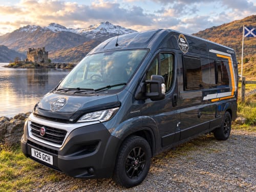 Black Fiat Ducato 4-berth campervan parked by Scottish loch with castle ruins and snow-capped mountains, Glasgow Camper Hire
