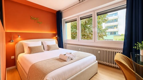 Modern bedroom at Mattbnb Luxembourg with vibrant orange accent wall, queen bed with white linens, large city windows, and