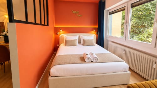 Bright orange bedroom at Mattbnb in Luxembourg Limpertberg with white bed, rolled towels, and city view windows