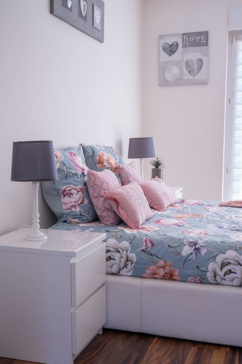 Acogedora habitación en la casa rural Les Ambrades de Colmar con ropa de cama floral, almohadas rosas y mesilla blanca con lámpara