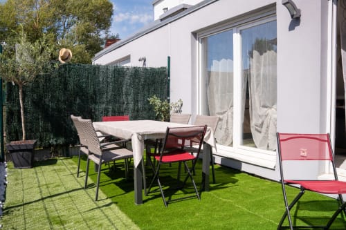 Soleada terraza de la casa rural Les Ambrades en Colmar con mesa de comedor, sillas rojas y seto verde