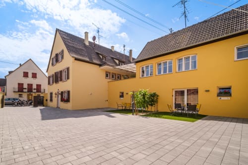 Patio colorido del hotel Les Ambrades en Sigolsheim, Alsacia, con edificios amarillos y crema con terraza