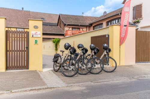 Cuatro bicicletas con cascos estacionadas en el hotel-cottage Les Ambrades en Sigolsheim, Francia, frente a la fachada amarilla del edificio
