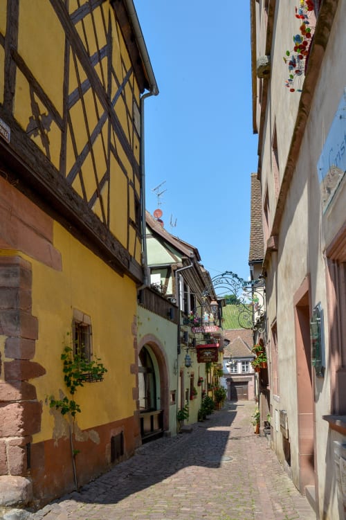 Encantadora calle medieval empedrada de Riquewihr con edificio amarillo de entramado de madera y flores coloridas, hotel Les Ambrades
