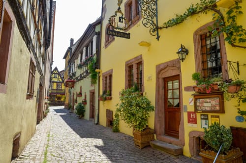 Pintoresco callejón de adoquines en Riquewihr con edificios de paredes amarillas, puertas de madera, hiedra y señalética tradicional