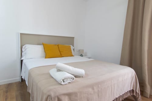 Camera moderna negli Apartamentos El Sol II con letto bianco, cuscini gialli, biancheria beige e asciugamani arrotolati a Tossa de Mar