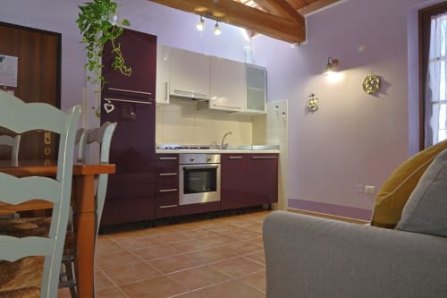 Zona soggiorno dell'appartamento Malva con pareti viola, cucina bordeaux, tavolo da pranzo e divano grigio presso La Casa dei Tigli, Cannara
