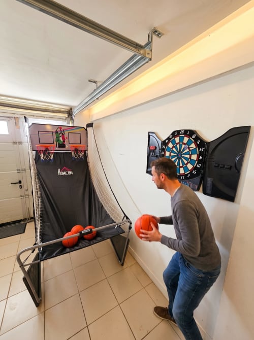 Homme jouant aux fléchettes dans la salle de loisirs du cottage Vindihouses, avec jeu de basketball et équipements de divertissement visibles