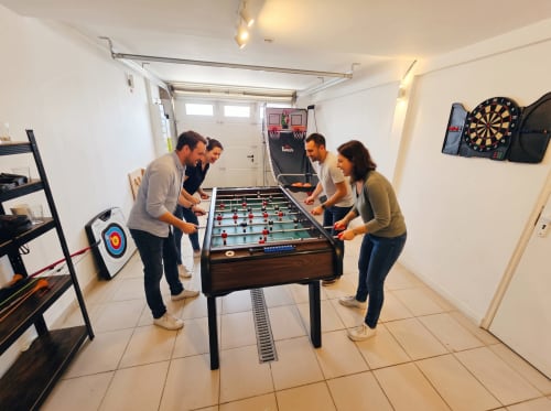 Cinq invités jouent au baby-foot dans la salle de jeux de la maison Vindihouses, avec fléchettes et jeux d'arcade visibles