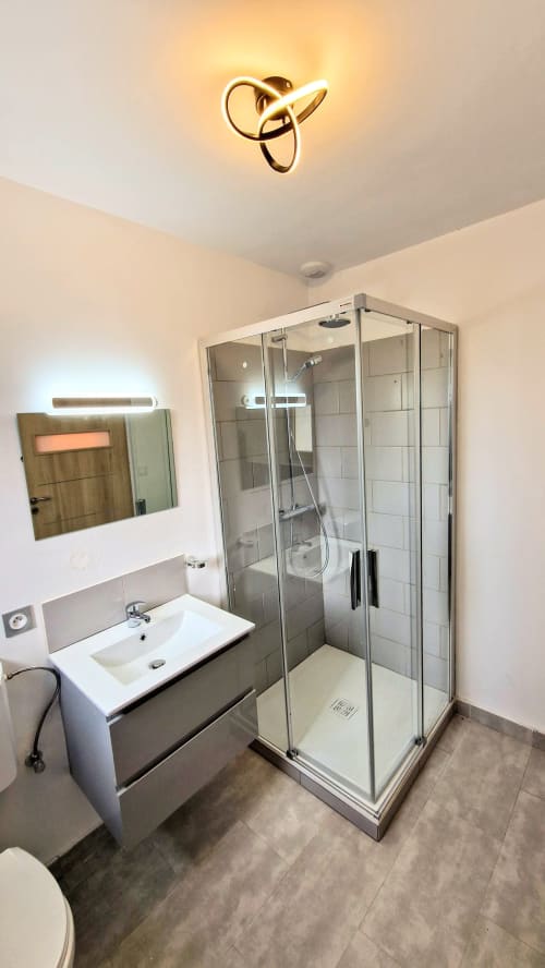 Salle de bains moderne du gîte Magnien avec douche vitrée, lavabo blanc et luminaire de plafond
