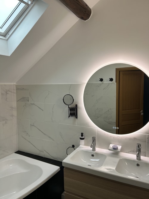 Salle de bains moderne au Domaine Chapelle : carrelage en marbre, miroir rond lumineux, double vasque et plafond mansardé avec puits de lumière