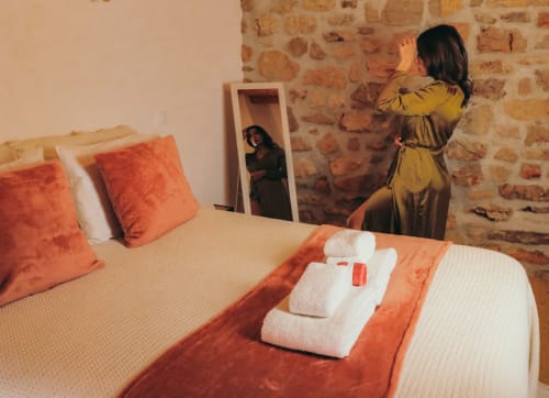 Quarto em Recantos D'Almerinda com paredes de pedra, acentos laranja, toalhas dobradas na cama, mulher em roupão amarelo olhando para o espelho